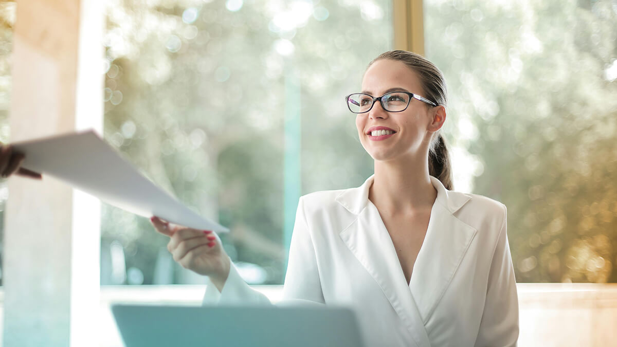 A woman in an office space smiling and exchanging papers with someone