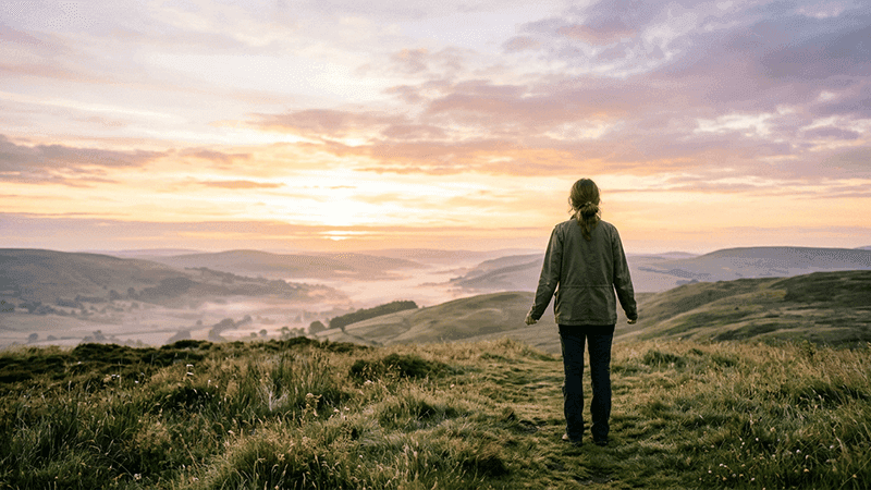 A woman watches the sun rise behind scenic rolling hills.